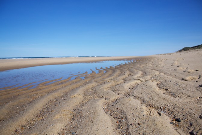 Close up of beach sand, with wavy pattern from waves, with ocean in background.