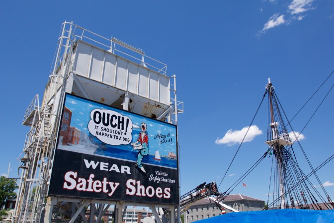 Billboard on building in shipyard. Text says "Ouch! It shouldn't happen to a dog: Wear Safety Shoes: Visit the Safety Shoe Store" and shows a sailor holding his foot, in pain.