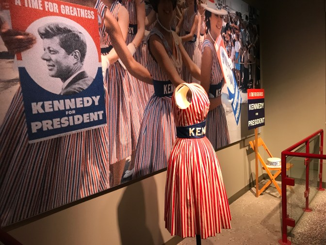 Display with photo of women wearing red and blue dresses with the word KENNEDY on a sash on them. A dress is on display in front of the photo.