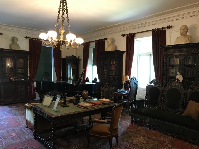 Library of the Longfellow House. A desk is in the foreground, a piano is in the background, and numerous bookshelves line the walls.