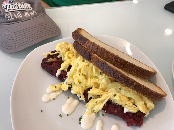 Eggs, hash, and toast, on a plate. A Patriots cap is in the background.