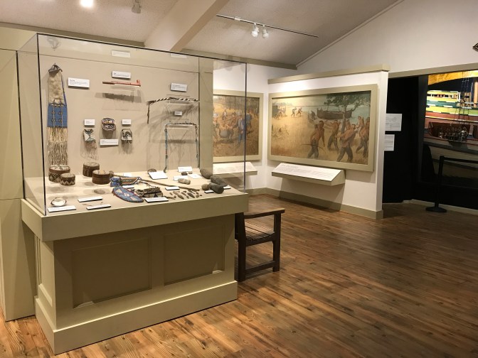 In the foreground, a display of Native American jewelry and other items. In the backgrounds mural depicting the first contact with Native Americans by the Pilgrims.