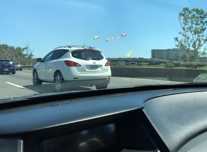 Kites flying by the side of the road. View through the front windshield of a car.