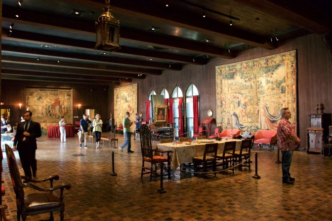 The Tapestry Room. Tapestries hang on the walls, a dinner table with chairs in in the center of the room. People are milling about the room.