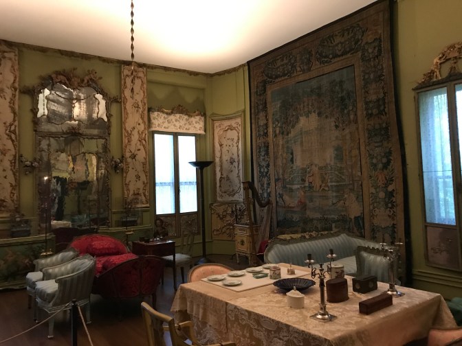 Little Salon. A tapestry hangs on the wall. A table is decorated with small plates. Chairs and benches surround the room.