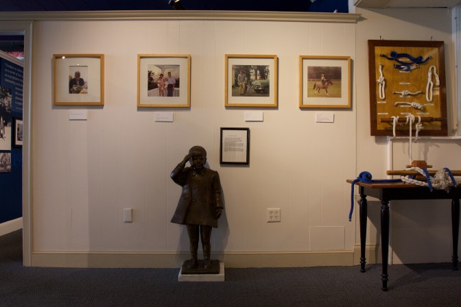 Display of photos of President Kennedy, nautical knots, and a statue of his son standing at attention.