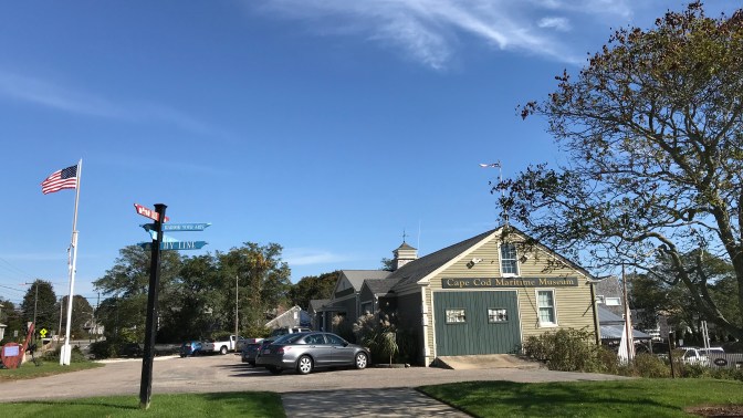 Cape Cod Maritime Museum. Cars are parked in the lot in front of the museum, and a flag flies on the left of the image.