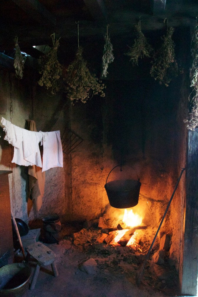 Pot smoking over an open flame. Herbs dry on the top of the hearth. Towels dry nearby.