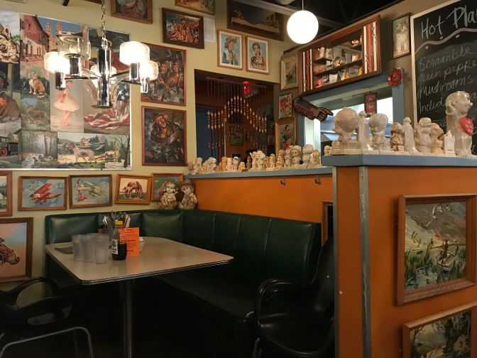Diner booth, with small porcelain dolls on the shelves behind the table. Paintings adorn the walls.