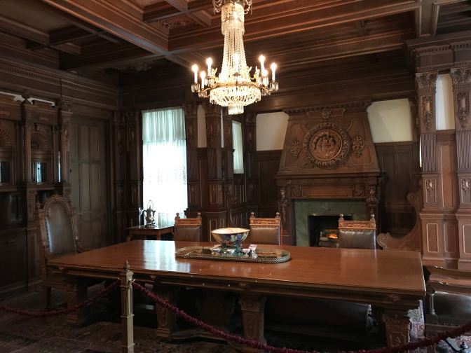 Dining room, with large wood table in the center, and a chandelier hanging above the table. A fireplace is in the background.