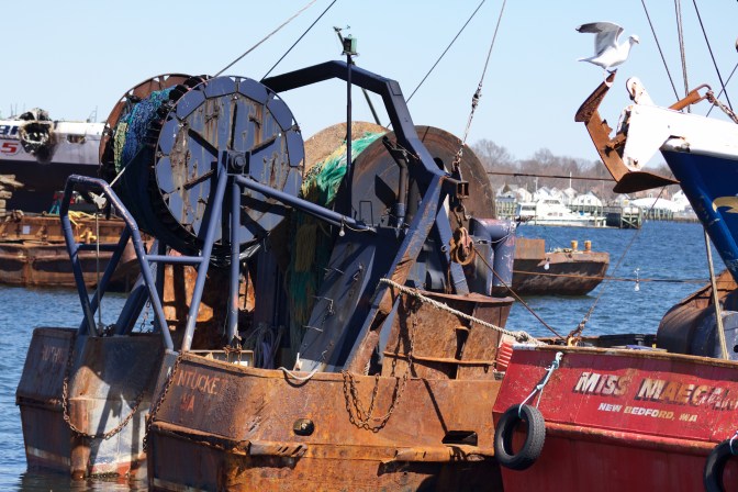 Stern of fishing boats. Boat in center of frame is heavily rusted.