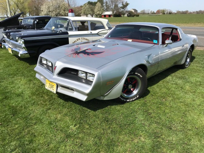 Silver Pontiac Firebird next to old police cruiser.