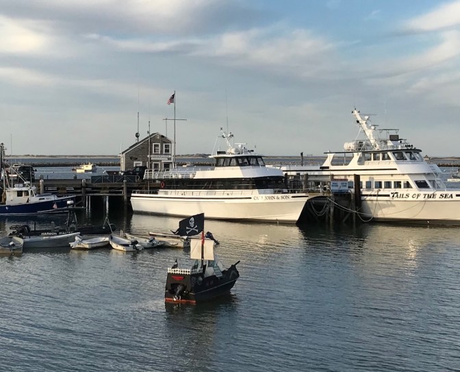 Plymouth Harbor, with a small pirate ship model in the water in front of two larger vessels.