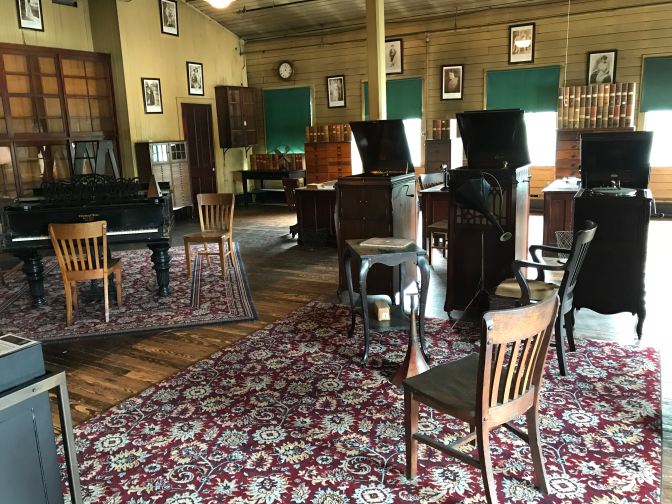 Three phonograph players in the middle of a large room. A piano is to the left of the players.
