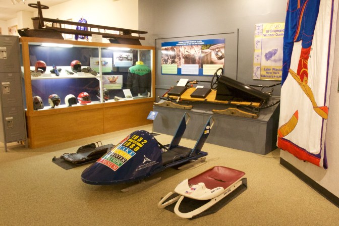 Interior of the Lake Placid Olympic Museum. A skeleton, luge, and bobsled are on the floor. A cutaway of a wooden bobsled is on the wall, along with a display case of bobsled gear including helmets and jackets.