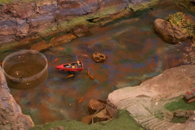 Miniature fishing pond with two model figures inside a model rowboat. Live fish swim in the water.