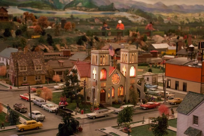 Miniature church, surrounded by cars parked on the street.
