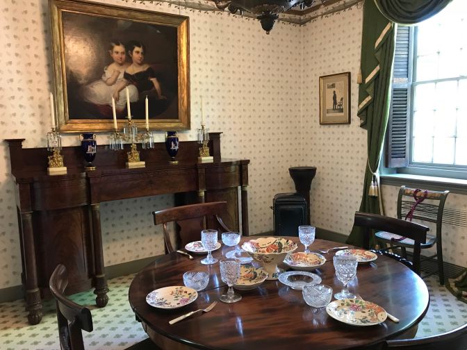 Empire Dining Room. A large painting of two girls is on the wall. A table set for four places is in the foreground.