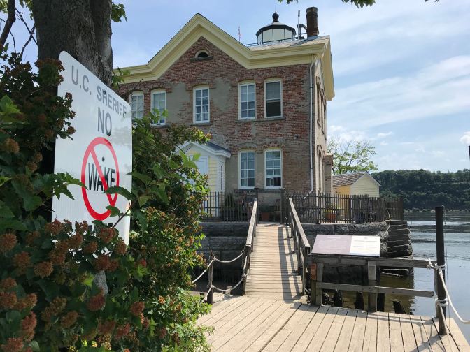 View of Saugerties Lighthouse with a wooden footbridge connecting the lighthouse to main land. A sign in the foreground says UC SHERIFF NO WAKE.