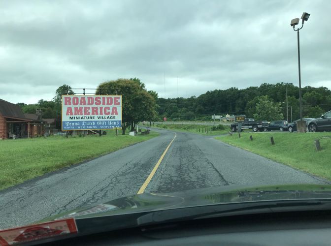 Two lane road. A sign on the left of the road says ROADSIDE AMERICA MINIATURE VILLAGE - DUTCH GIFT HAUS- GIFTS HEX SIGNS - SOUVENIRS