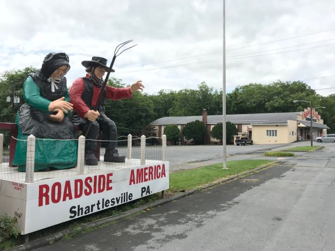 Statues of Amish man and woman seated on a plinth, waving at the traffic.