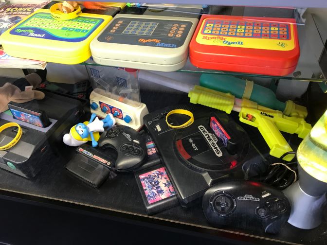 Display case including a Speak and Spell, a super soaker, and a Sega Genesis.