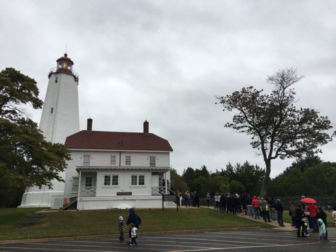 Exterior of Sandy Hook Lighthouse, with a line of people extending out to the parking lot from the rear porch.
