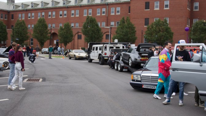 View of cars, with spectators dressed in 1980s and 1990s attire.