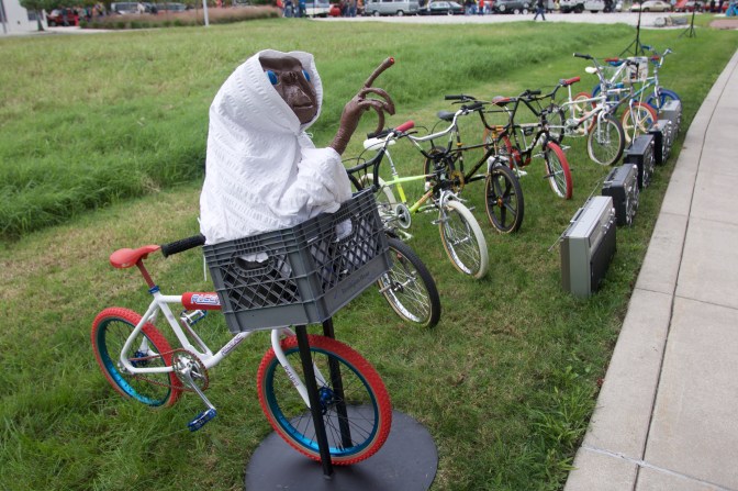 Model of E.T. in a bicycle basket, in front of a row of children's bikes from the 80's and boombox stereos.