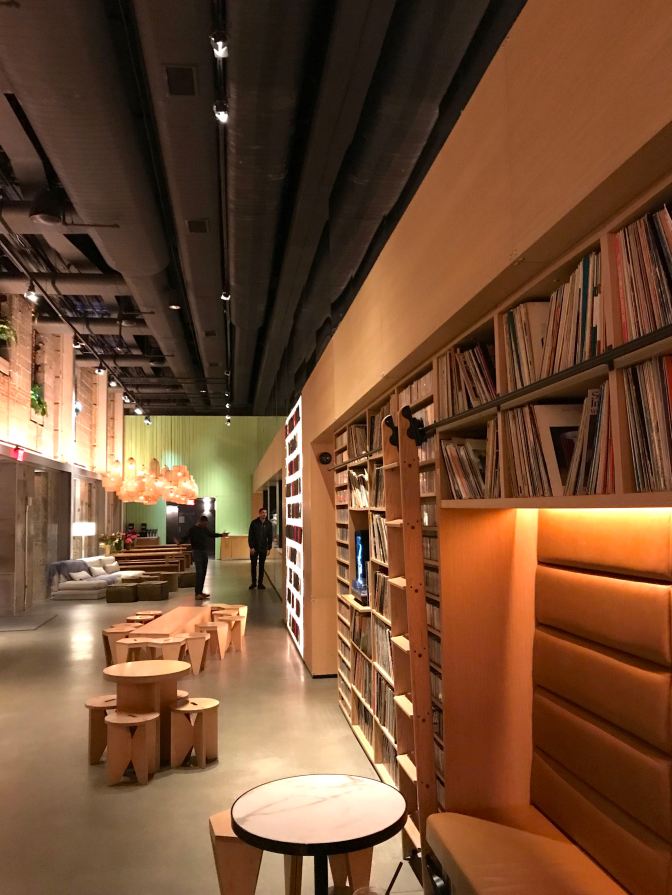 Hotel lobby. Small wooden chairs and tables are in the middle of the floor. Two men are in the background. Shelves filled with LP records and cassettes line the walls.