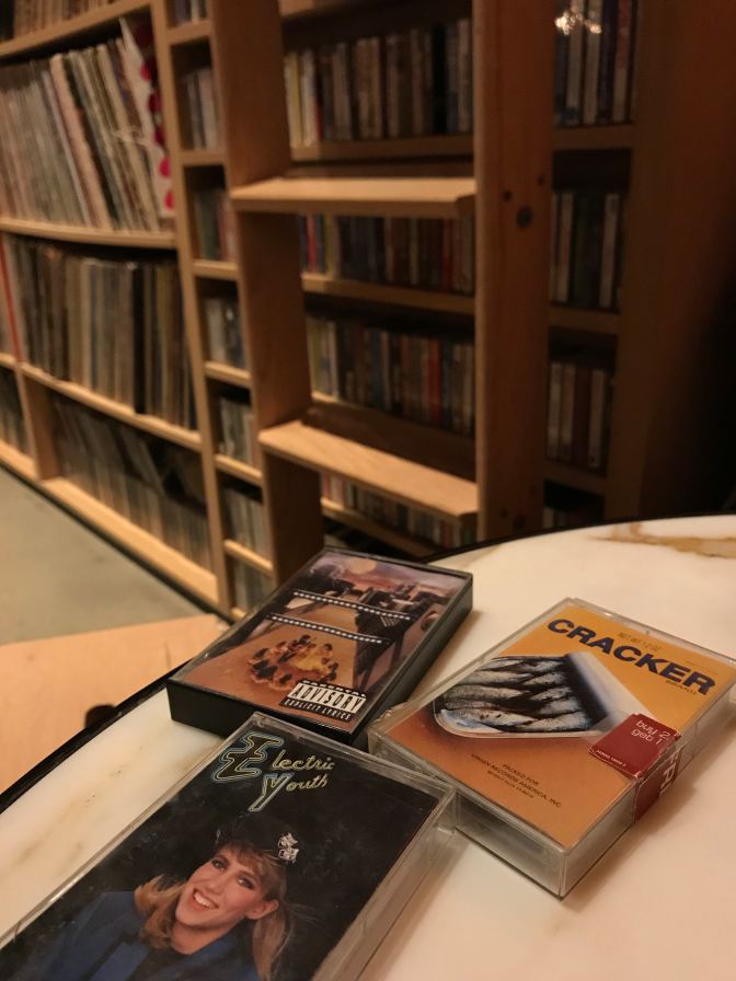 Cassette tapes on table, with records and cassettes on shelves in background.