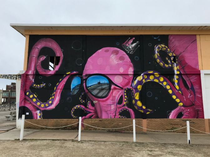 Mural of pink Octopus on black background, wearing a crown and sunglasses. The boardwalk is reflected in the sunglasses.