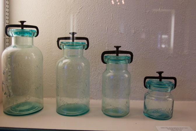Four mason jars of varying heights in a glass case.