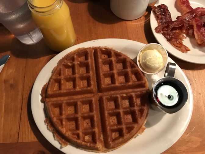 Waffle on plate with butter and maple syrup. Bacon is on another plate, and a glass of orange juice is at the top left of the image.