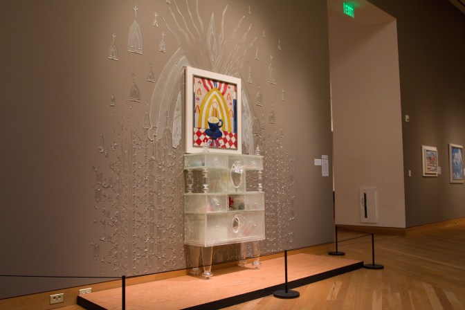 Cabinet and painting in glass along wall in museum. Shards of glass decorate the wall beside it.