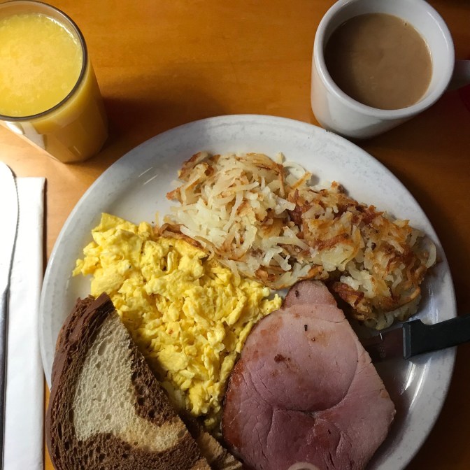 Plate with scrambled eggs, ham, hashed browns, and rye toast. A cup of coffee and a glass of orange juice is on the table.