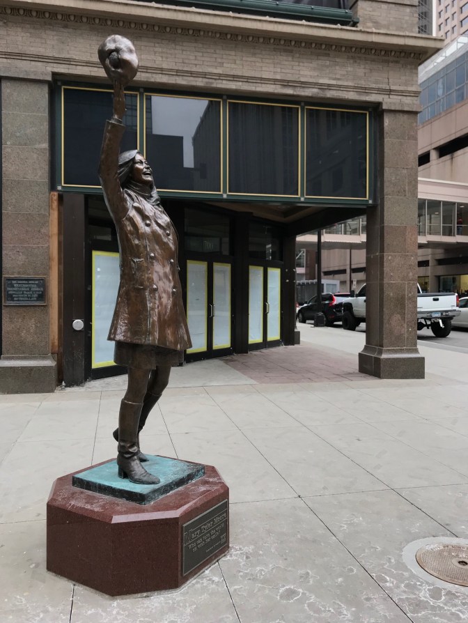 Statue of Mary Tyler Moore on pedestal in front of brick building.