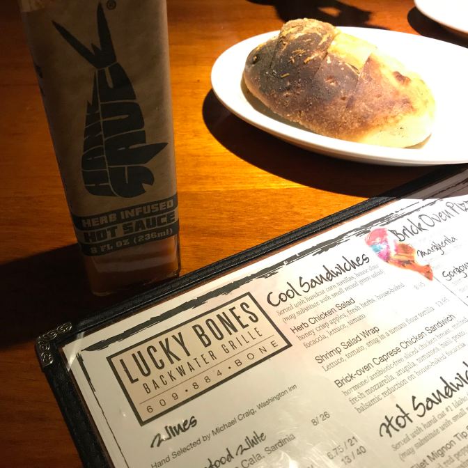 Menu at Lucky Bones Backwater Grille restaurant with a roll on a white plate and a bottle of hot sauce in the background.
