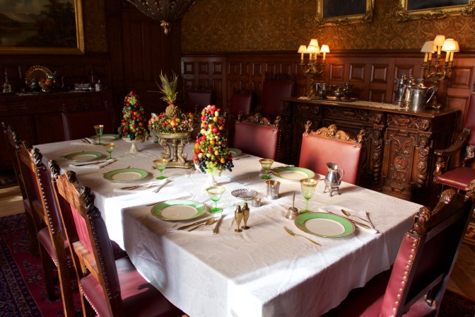 Dining room of the castle. The large rectangular table has places set for 8 people, with gold flatware and white plates with gold rims. There are three centerpieces made of fruit, and a sideboard holds water tankards and lights.