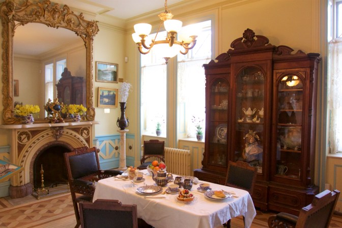 Breakfast room, with a table set for four, a large mirror over the fireplace, and a sideboard filled with tea sets and plate sets.