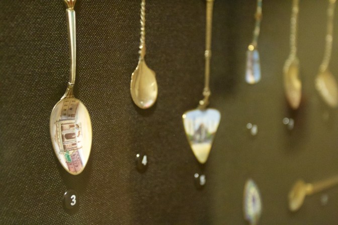 8 spoons on display in a case. The spoon on the left has an engraving of a mosque.