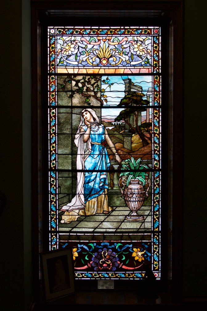 Stained glass window with woman standing on patio, a plant in a ceramic vase beside her.