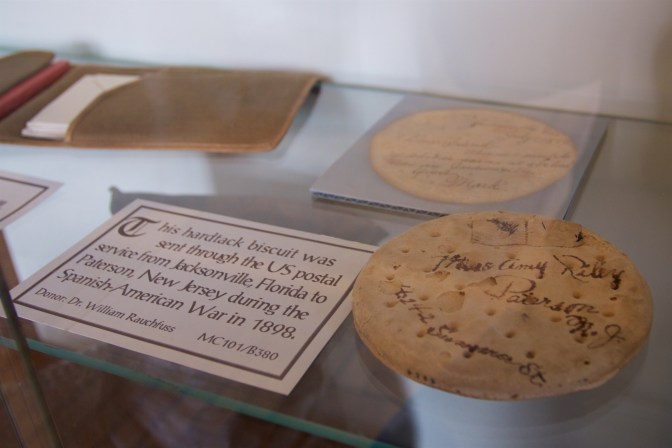 Hardtack cracker on a glass shelf. A sign next to it says THIS HARDTACK BISCUIT WAS SENT THROUGH THE US POSTAL SERVICE FROM JACKSONVILLE FLORIDA TO PATERSON, NEW JERSEY DURING THE SPANISH-AMERICAN WAR IN 1898.