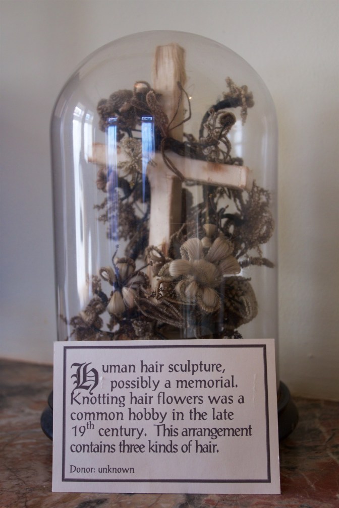 Cross with floral decorations under glass case. A sign in front of it says HUMAN HAIR SCULPTURE POSSIBLY A MEMORIAL. KNOTTING HAIR FLOWERS WAS A COMMON HOBBY IN THE LATE 19TH CENTURY. THIS ARRANGEMENT CONTAINS THREE KINDS OF HAIR. DONOR UNKNOWN.