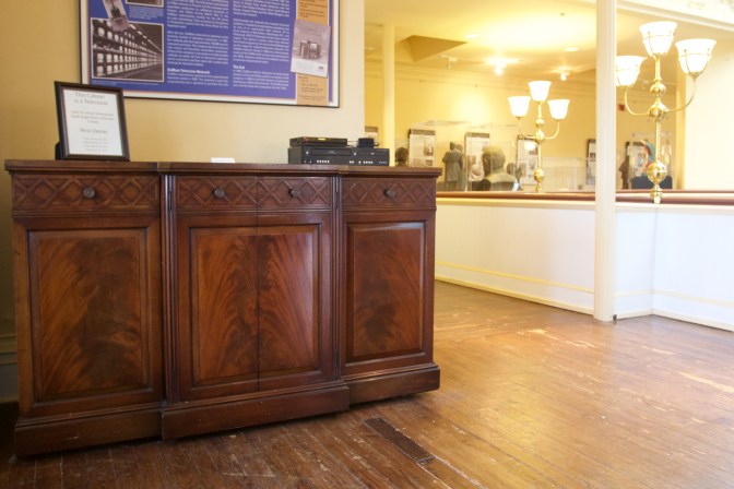 Large cabinet television on wooden floor. A sign is on top of it and another sign (both illegible) are on the wall. A small VCR and DVD player sits on the top of the TV.