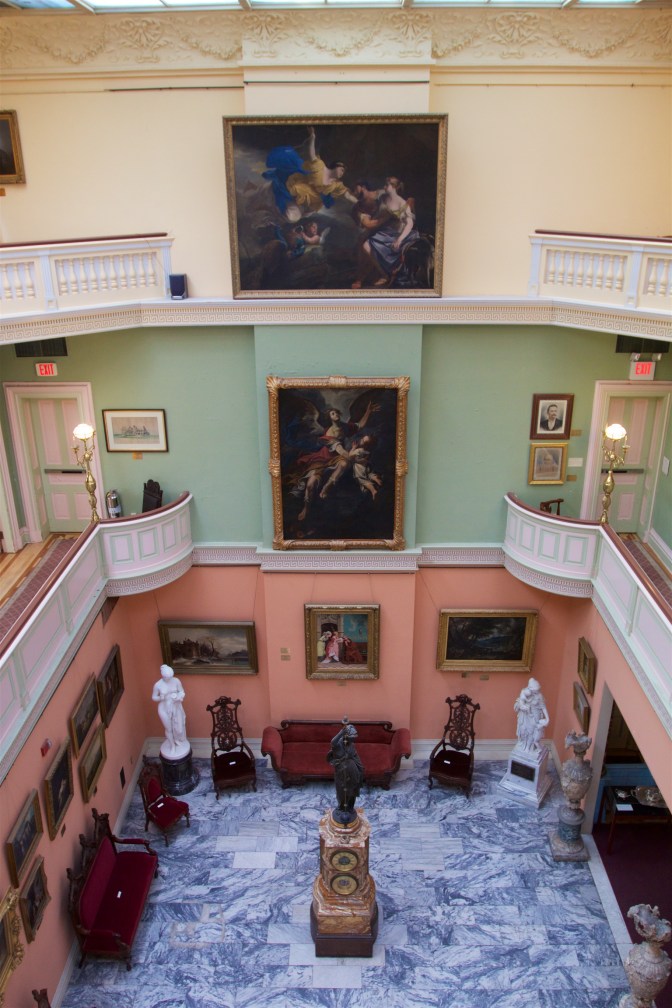 View of Grand Atrium from third floor. Paintings hang on walls, statues are on the first floor along with chairs and benches. The clock is in the center of the floor.