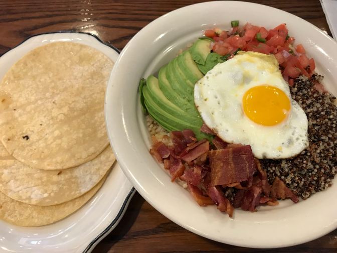 Plate on left has three corn tortillas. Plate on right has fried egg, quinoa, brown rice, tomatoes, avocado, and bacon.