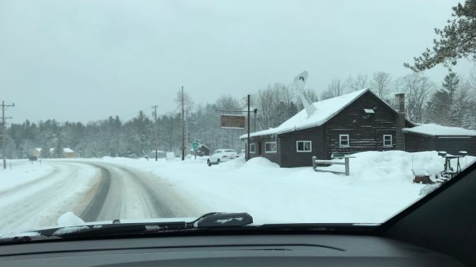 View of Wigwam Tavern on the side of a snow-covered road. View is from behind dashboard of Jeep.