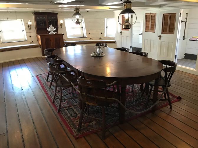 Captain's mess table, surrounded by eight chairs. White wood paneling surrounds the room.