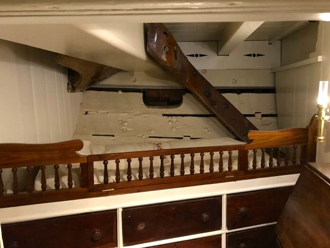 Chaplain's quarters, with a bed above a chest of drawers. A large beam in the ceiling is about two feet above the bed.